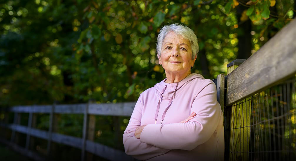 Smiling elderly woman leaning against a fence under the trees.